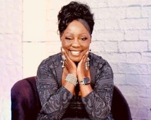woman with long black hair pulled up in a cute style. She's sitting in front of a white brick wall with her hands resting on her chin and smiling big. She's wearing a gray/black sweater with designs and lots of rings, a bracelet, and watch