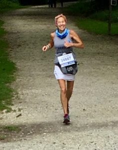 woman running on a trail in a gray tank and white running skirt, smiling, and enjoying nature