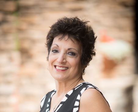 woman with short black hair turned to the side looking forward with a smile on her face. She's wearing a black tank-style dress with a white and black border. The background is blurred out