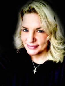 Headshot of a woman with medium blonde hair smiling and wearing a black shirt and a silver cross necklace